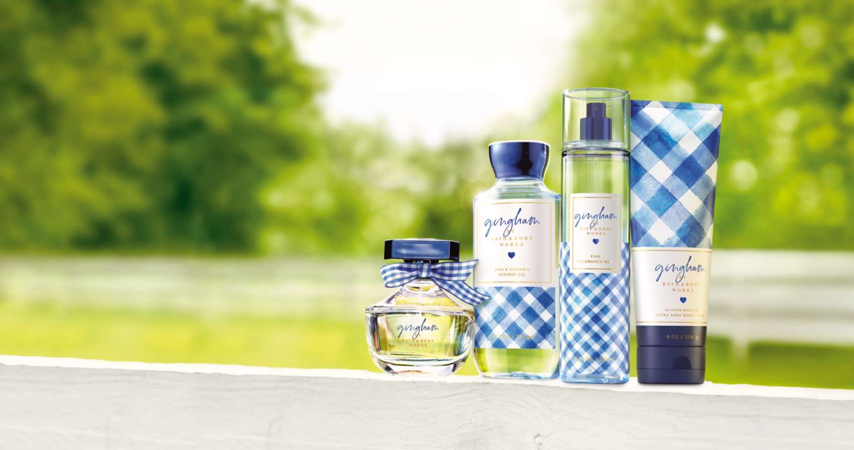 A countryside, tree-lined, white pickett fence with trees in the background and a gift set standing up on the fence in the foreground