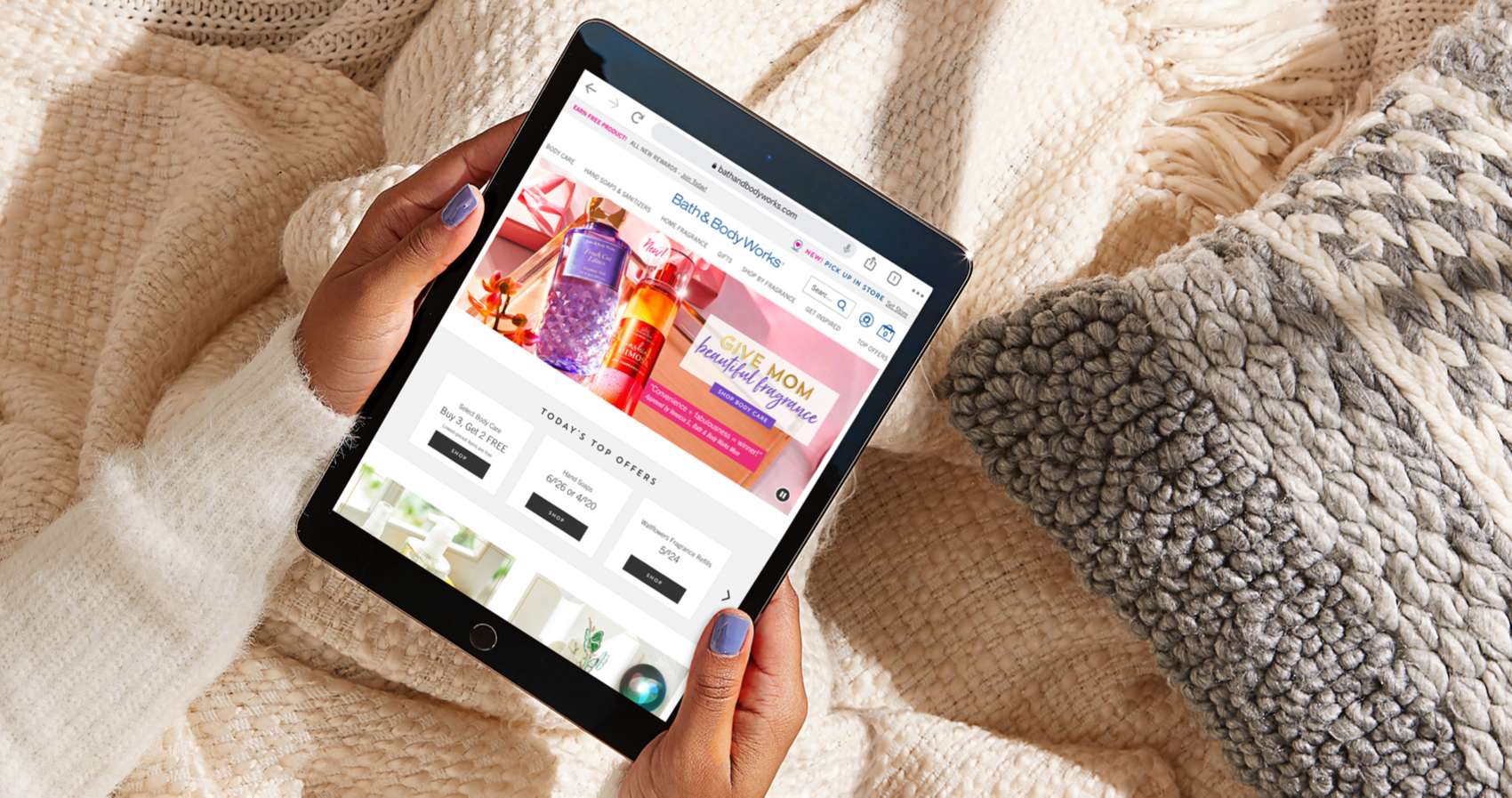 A person at home holding a smart tablet searching the Bath and Body Works website comfortably sitting on a bed of woven knits and blankets.