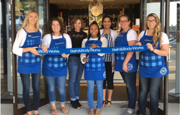 A diverse team of Bath and Body Works employees wearing their uniformed blue aprons holding a blue ribbon together in front of a new store location.
