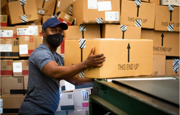 A shipping facility stacked with brown boxes from left to right with a make employee wearing a mask and a hat as he handles a taped box.